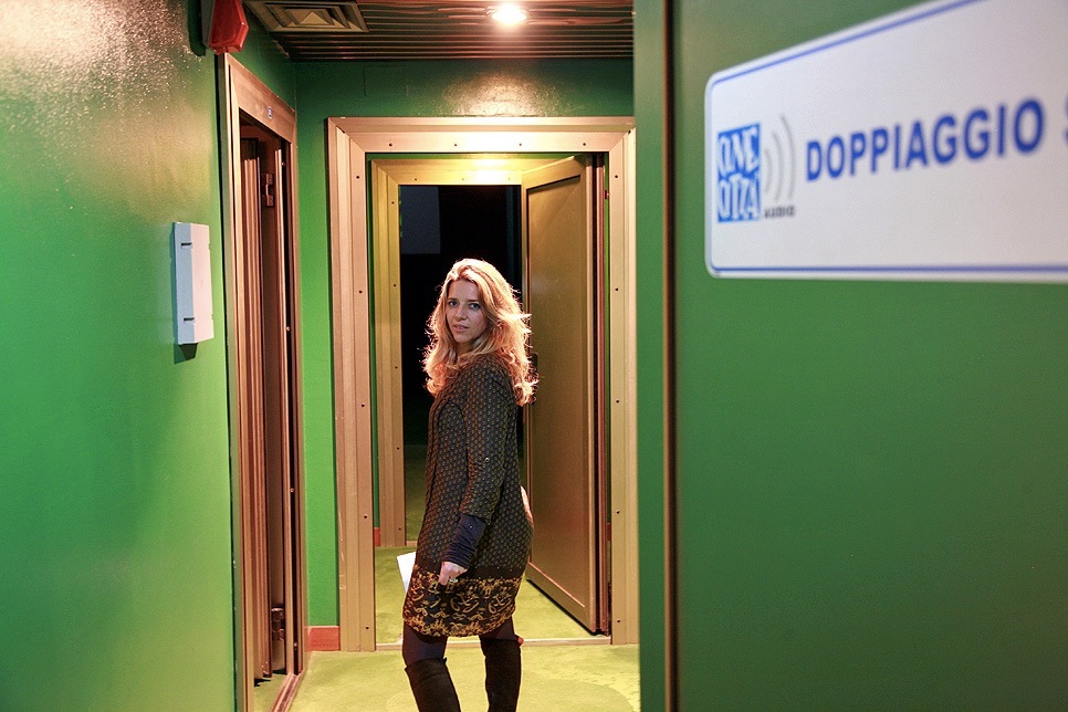 Woman with reddish-blonde hair standing in a green-walled dubbing studio corridor labeled 'DOPPIAGGIO'