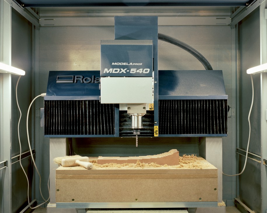 A Roland Modela Pro II MDX-540 CNC-milling machine with wood shavings and a carved wooden piece on its bed.