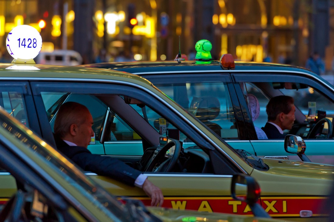 Tokyo taxi drivers wait in queue at night with illuminated rooftop signs and meters visible.
