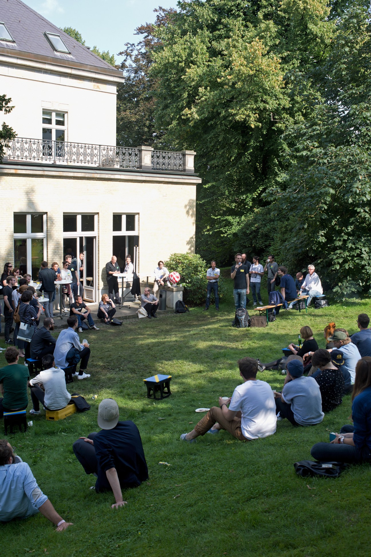Conference attendees sit on a manicured lawn in the garden of a white villa, listening to presentations near the building.