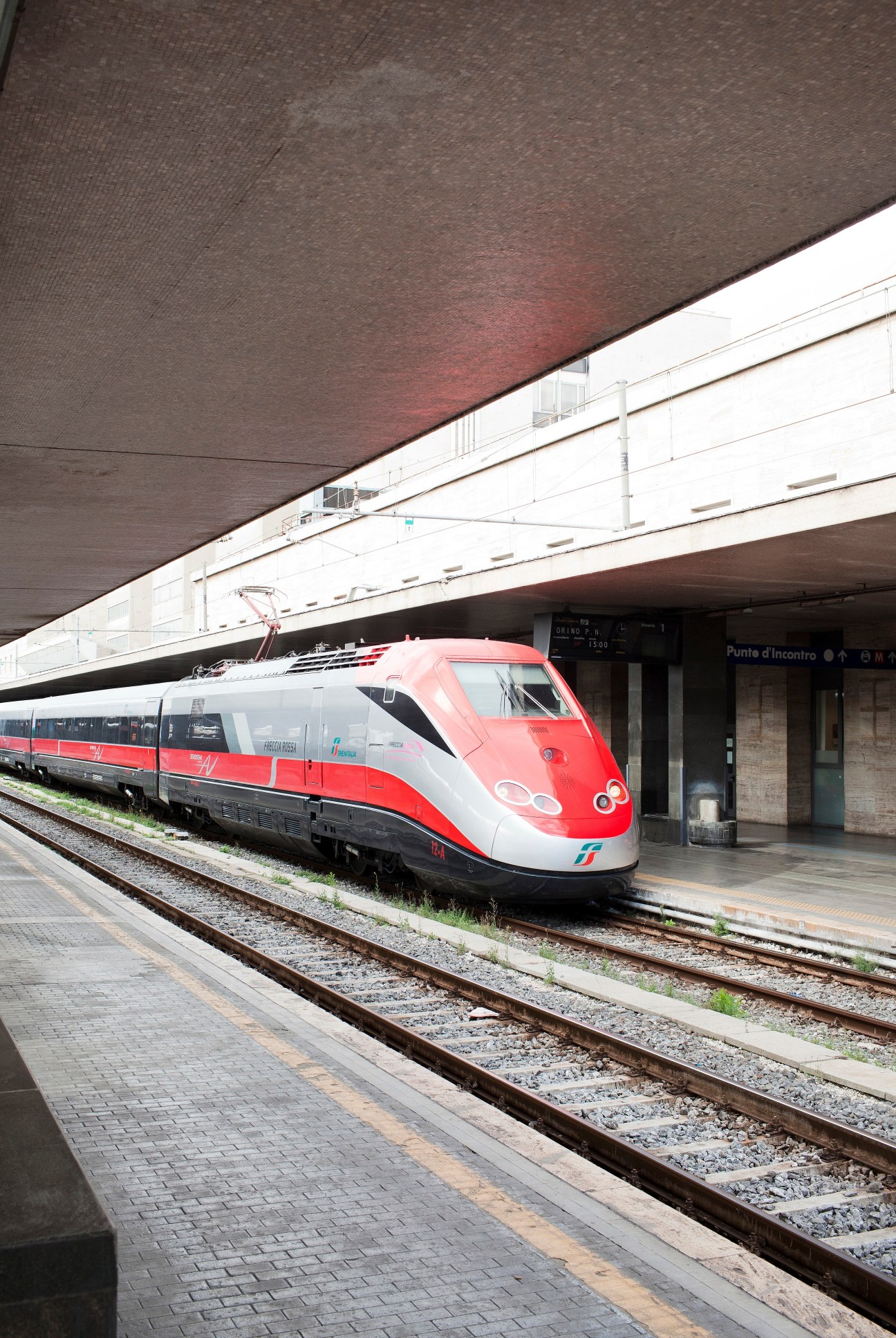 Red and white Frecciarossa high-speed train at platform under concrete overpass.