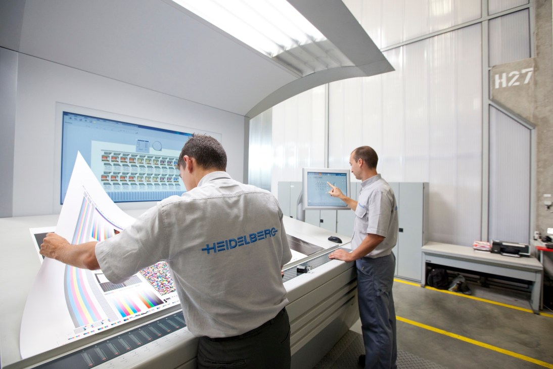 Two Heidelberg technicians inspect printed color samples at a quality control workstation.