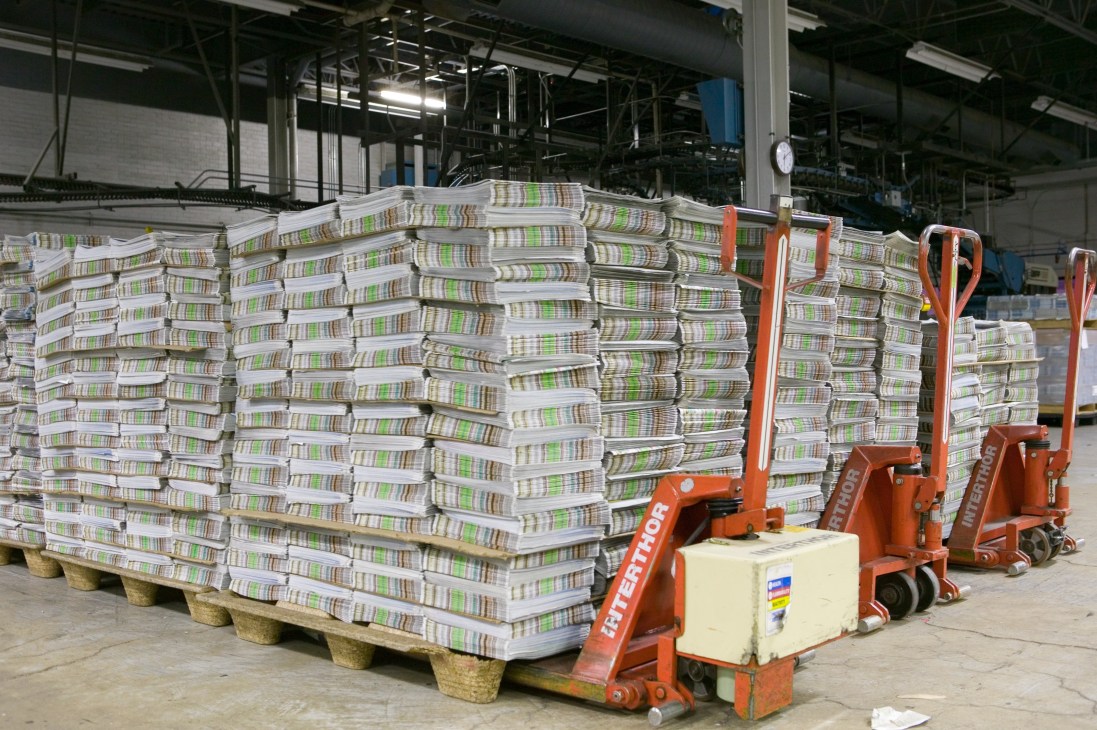Stacks of newspapers wrapped in plastic sit on pallets with red hand trucks in a warehouse.