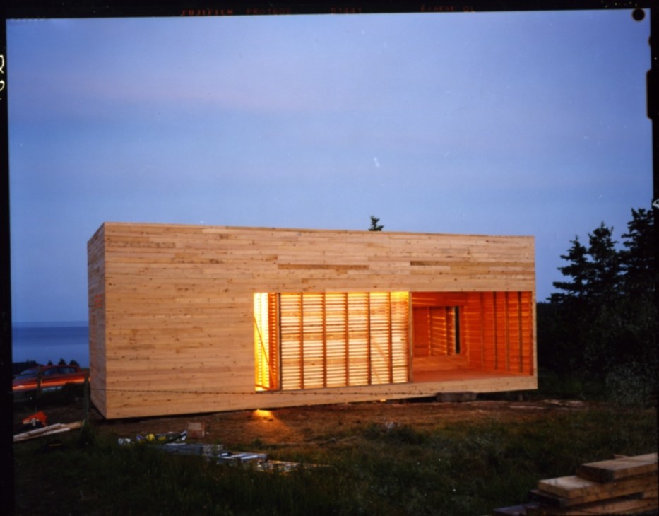 Wooden sleeping house glowing from interior lights at dusk with ocean visible in background.