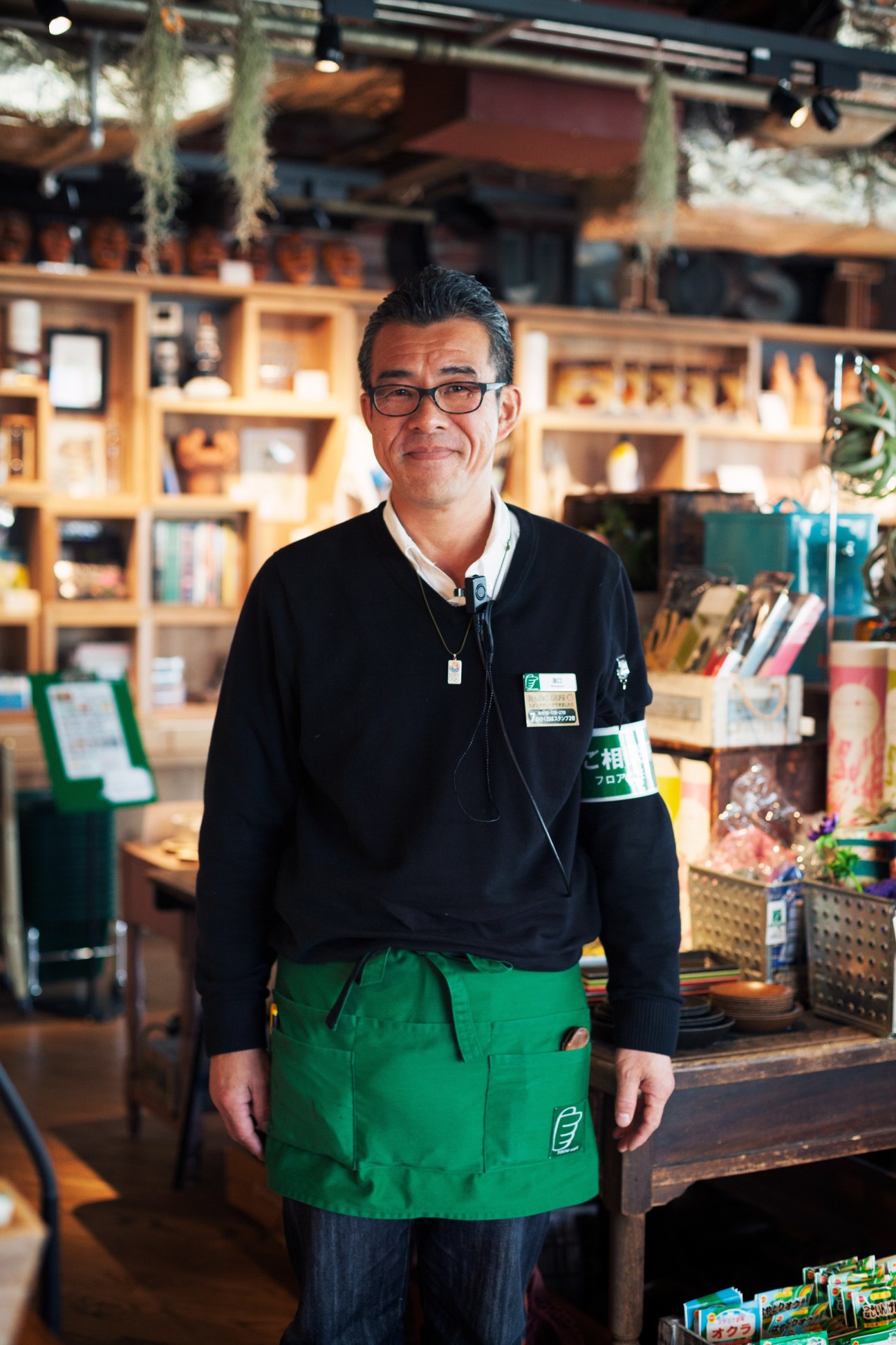 Floor manager Hiroshi Mizoguchi wearing Tokyu Hands green apron and staff badge in store with wooden shelving behind.