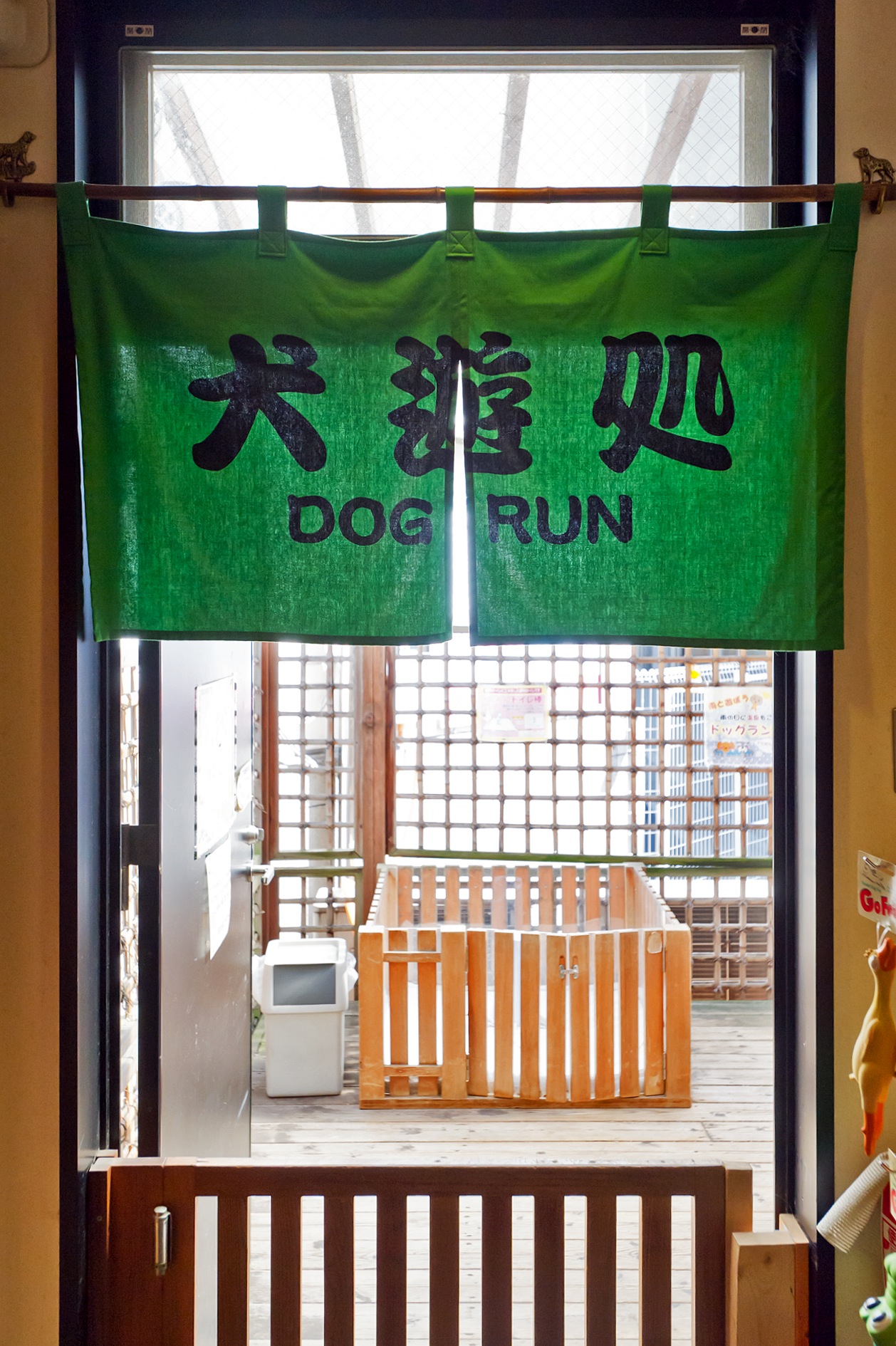 Green fabric banner with Japanese characters and 'DOG RUN' text hanging above wooden gate entrance to outdoor dog play area.