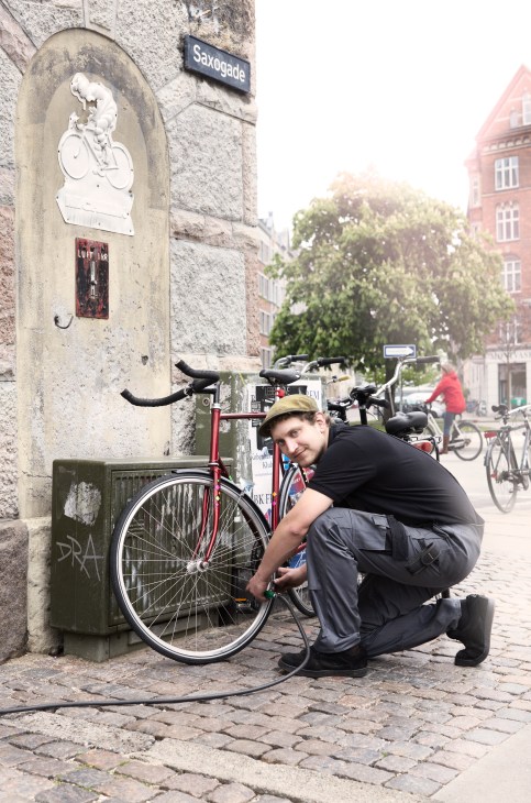 A man crouches beside a red bicycle at a Copenhagen street pump station near a Saxogade street sign.