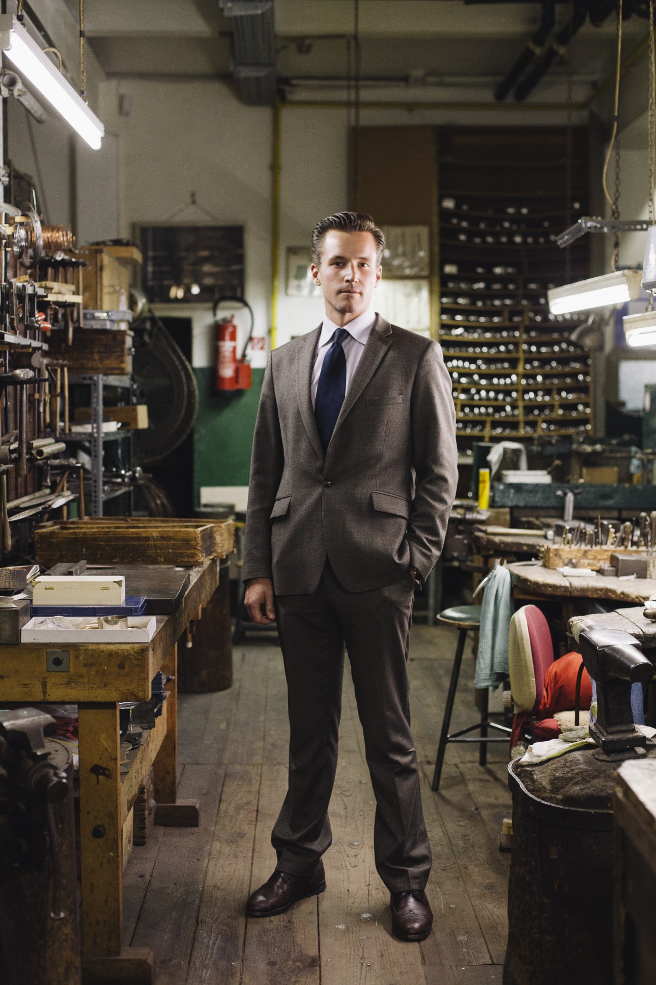 Man in grey suit standing in a workshop with leather goods and metalworking equipment visible in background.