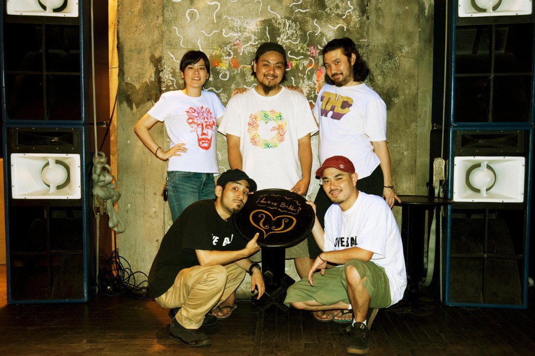 Five people posing in front of a weathered wall with decorative murals, holding a Love Ball Crew sign.