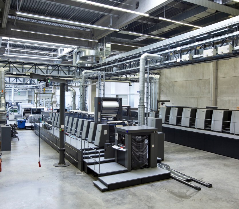 Industrial printing press machinery on factory floor with metal catwalk and overhead lighting systems.