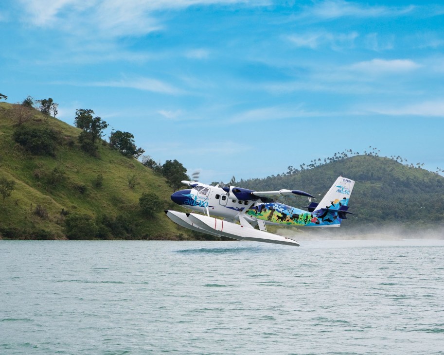 De Havilland Twin Otter seaplane landing on water with forested hills in background.