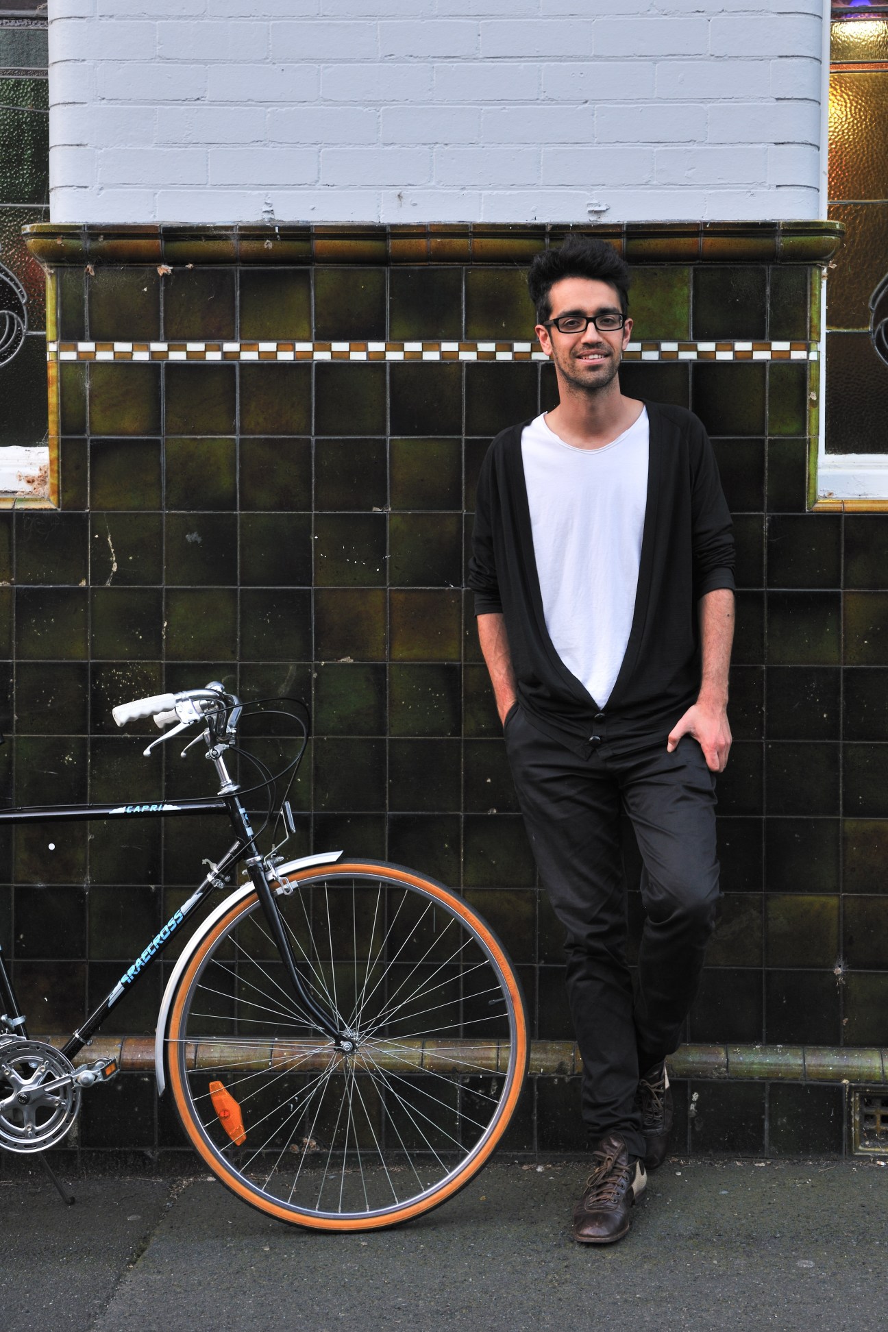 Man in black cardigan and white t-shirt standing with a vintage bicycle with orange accents against a tiled wall.
