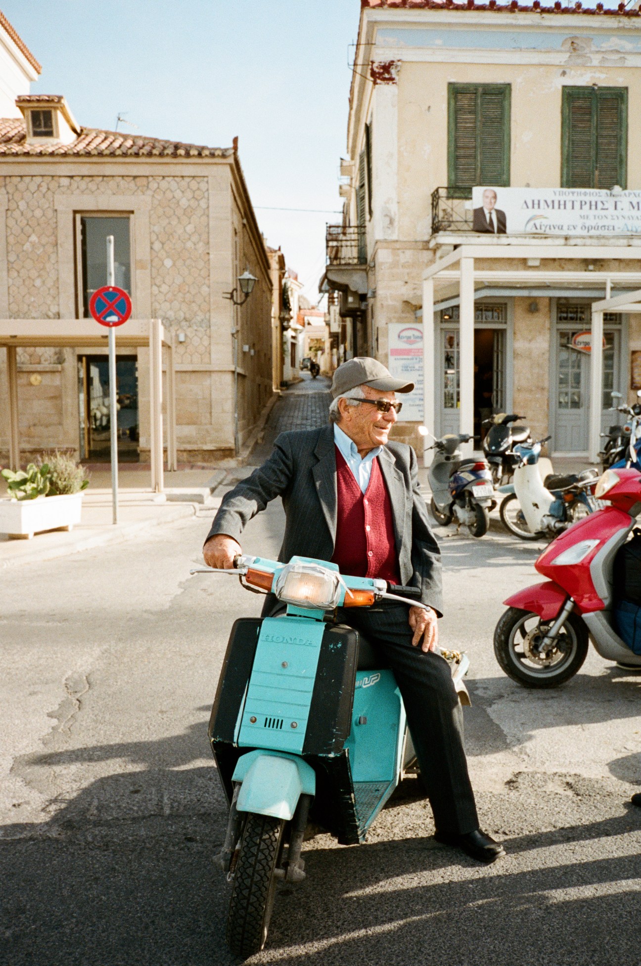 Elderly man riding a turquoise Vespa scooter on a narrow Aegina street lined with neoclassical buildings.