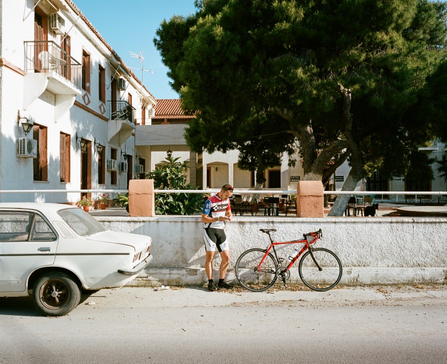 Man in cycling gear takes a break with his red bicycle on a quiet Aegina street lined with neo-classical buildings.