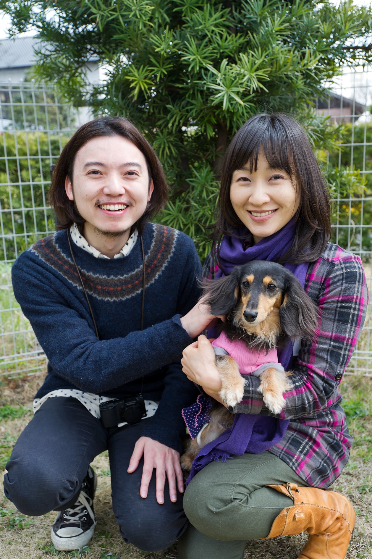 Two smiling women crouch beside a dachshund wearing a pink sweater in a garden setting.