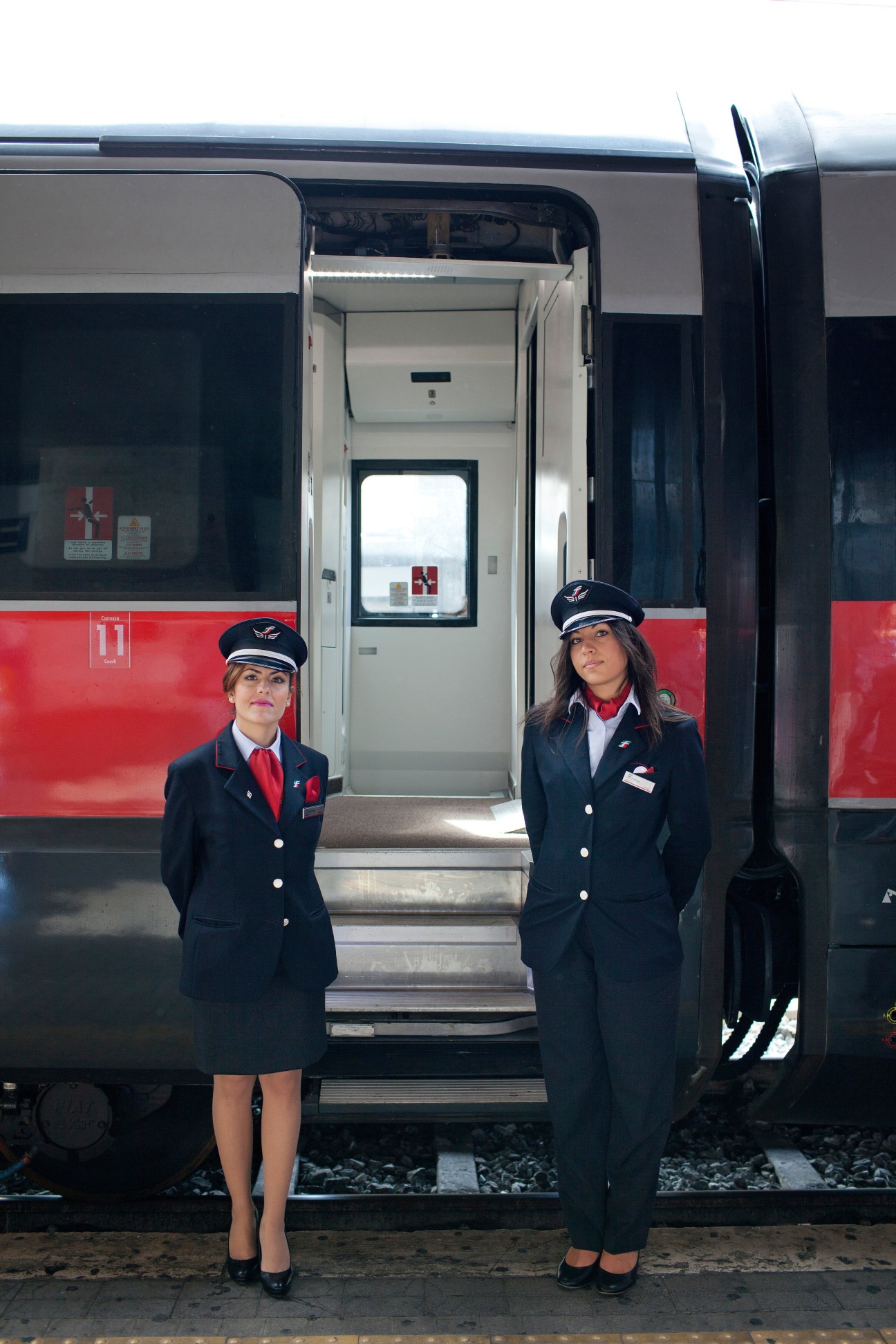 Two female train conductors in navy uniforms and red accents stand in front of an open Freccia train door.