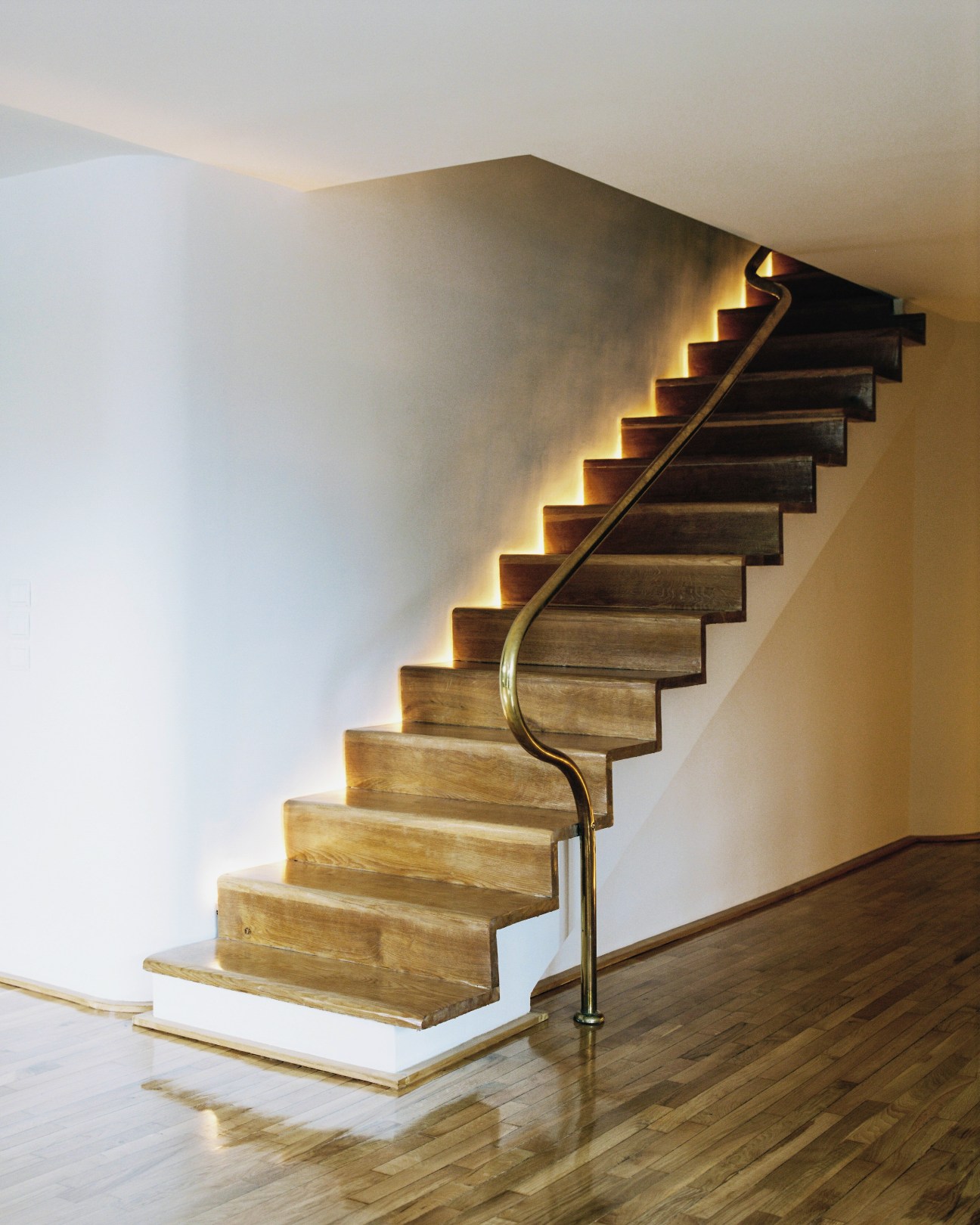 Modern wooden staircase with curved metal handrail and warm uplighting against white walls.