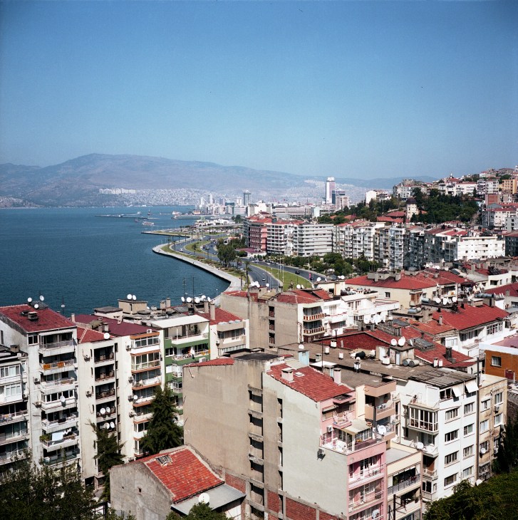 Aerial view of Izmir's waterfront with apartment buildings, bay, and mountains in the distance.