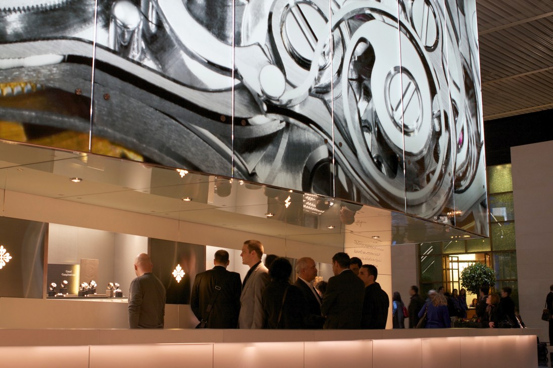 Visitors at the Patek Philippe stand at Basel watch fair with large watch mechanism display above.