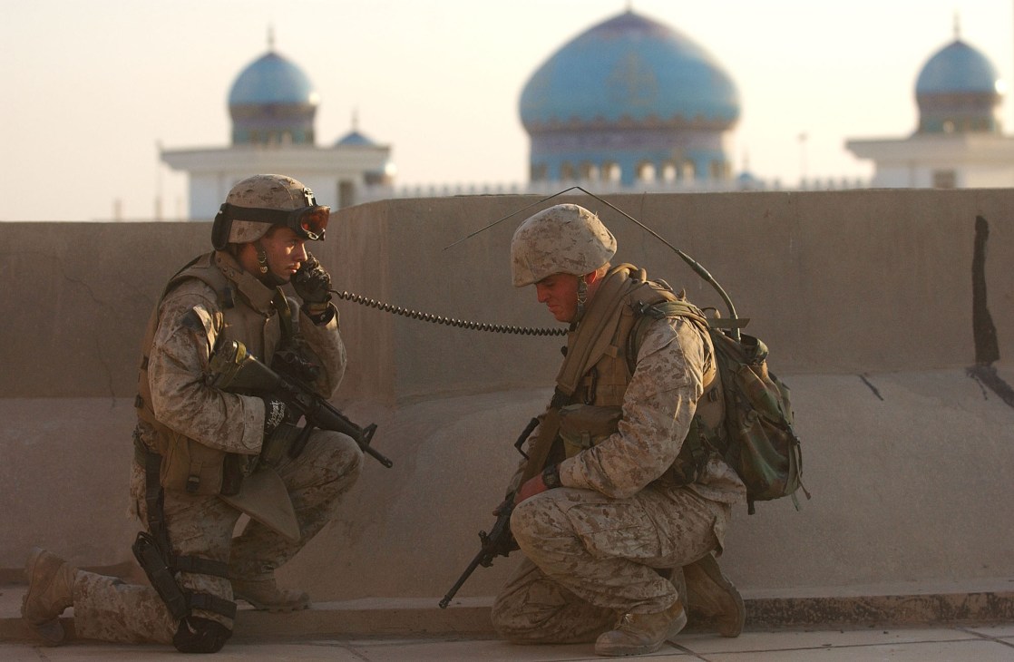 Two soldiers in desert camouflage uniforms communicate via radio headset while kneeling behind a concrete barrier with blue-domed buildings in the background.