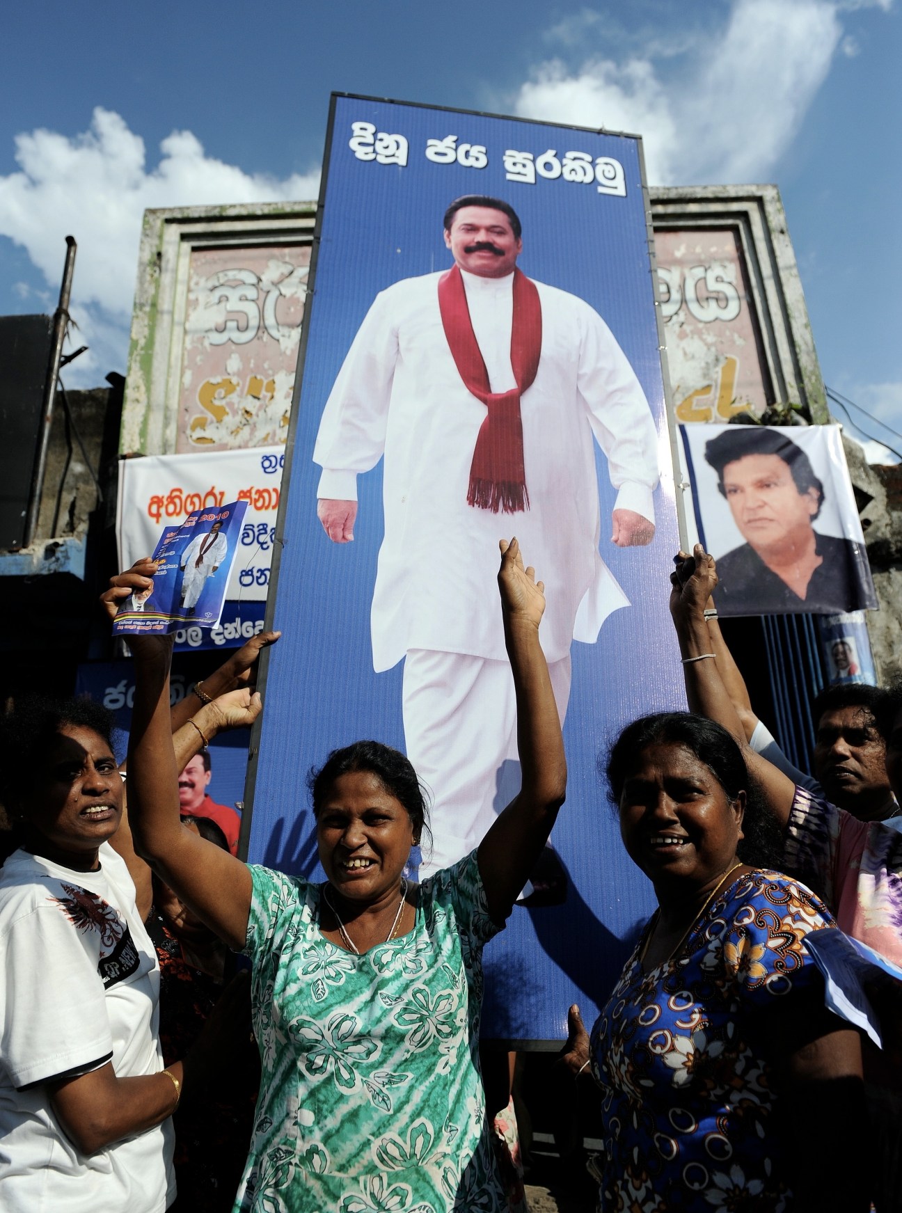 Supporters hold photographs and signs beneath a large blue billboard featuring Sri Lankan president Mahinda Rajapaksa in white clothing with a red scarf.