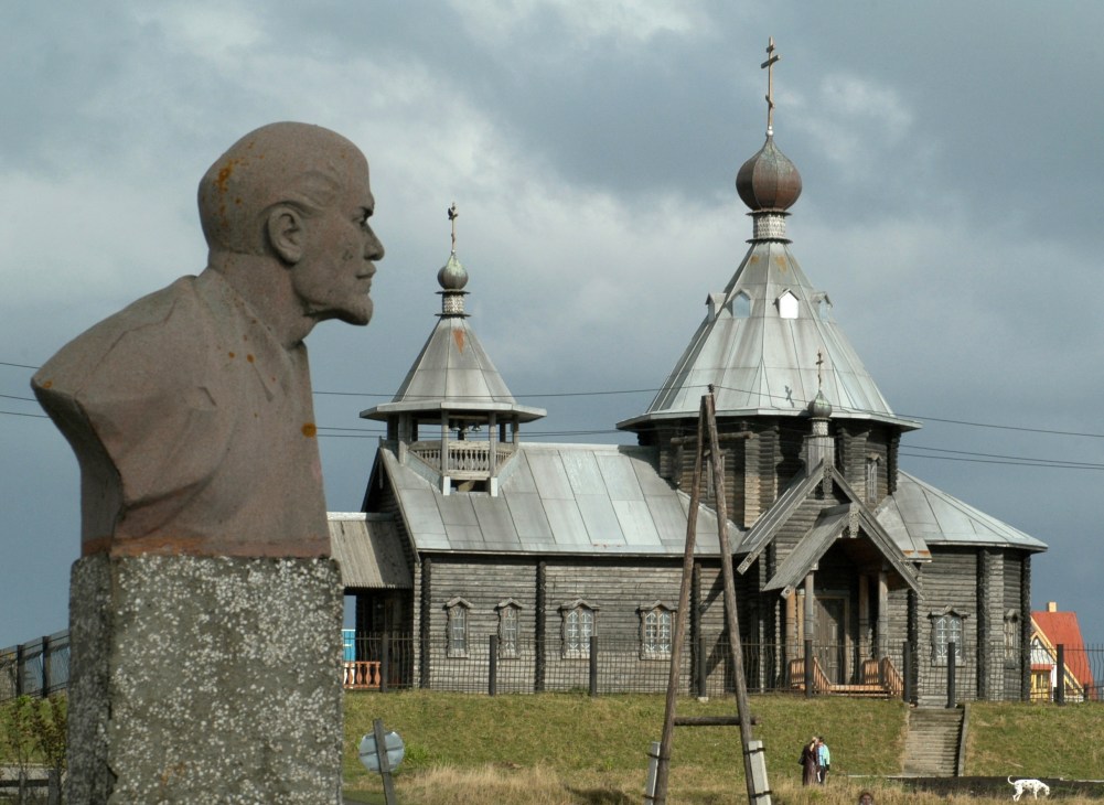 Stone bust sculpture facing a Russian Orthodox church with onion domes on an overcast day.