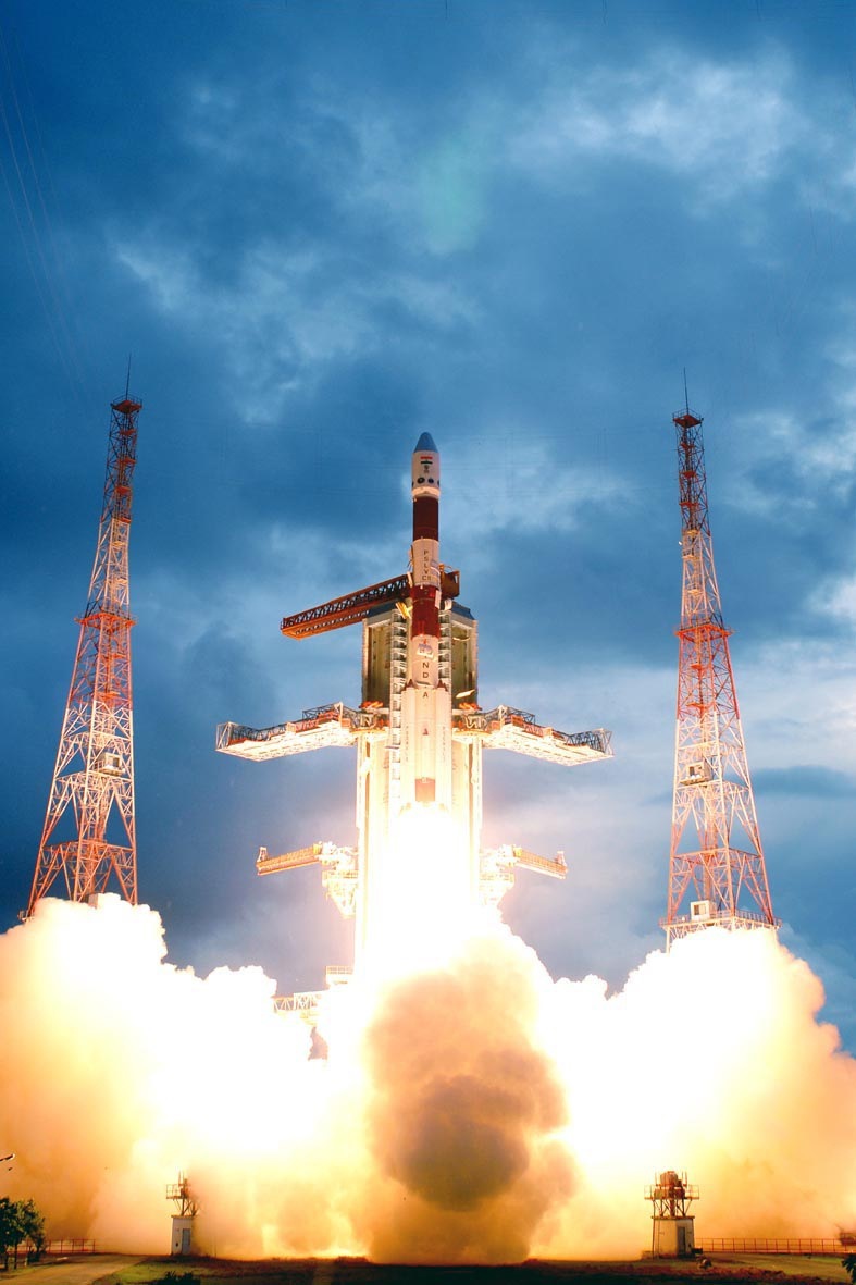 Indian space rocket launching with flames and smoke, flanked by red and white transmission towers.