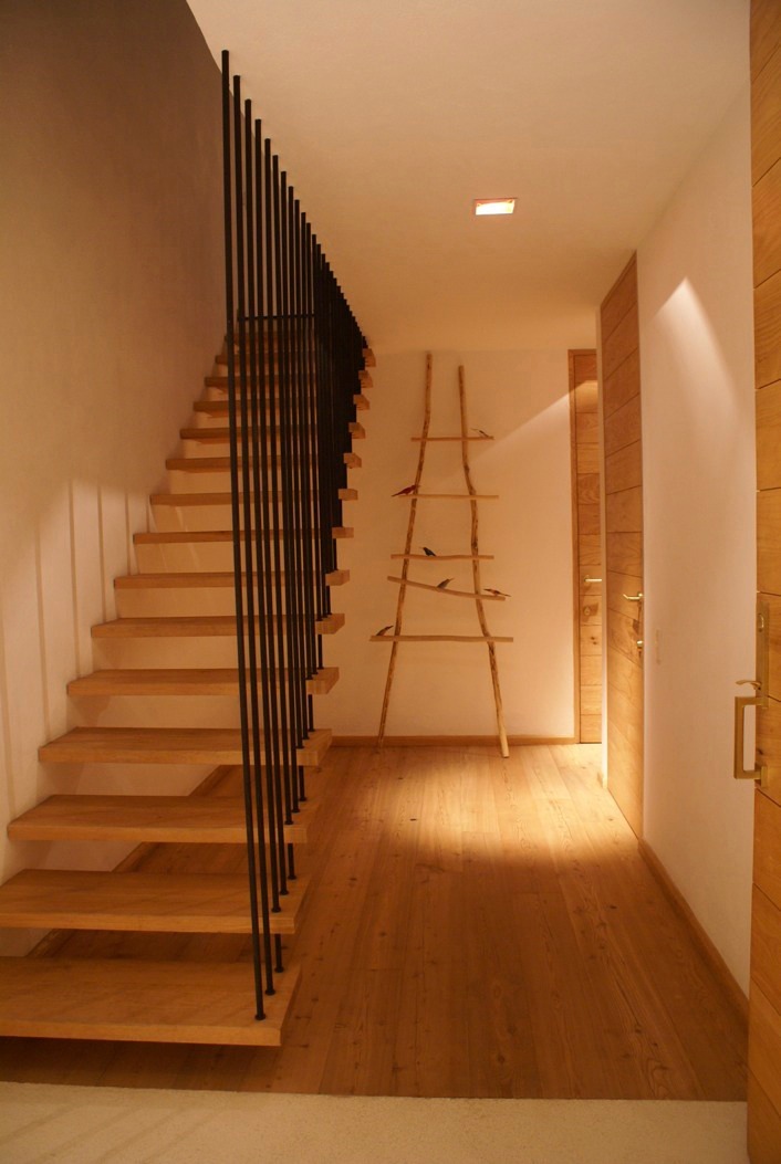 Wooden staircase with black metal railing in a light-filled interior hallway with wooden flooring.