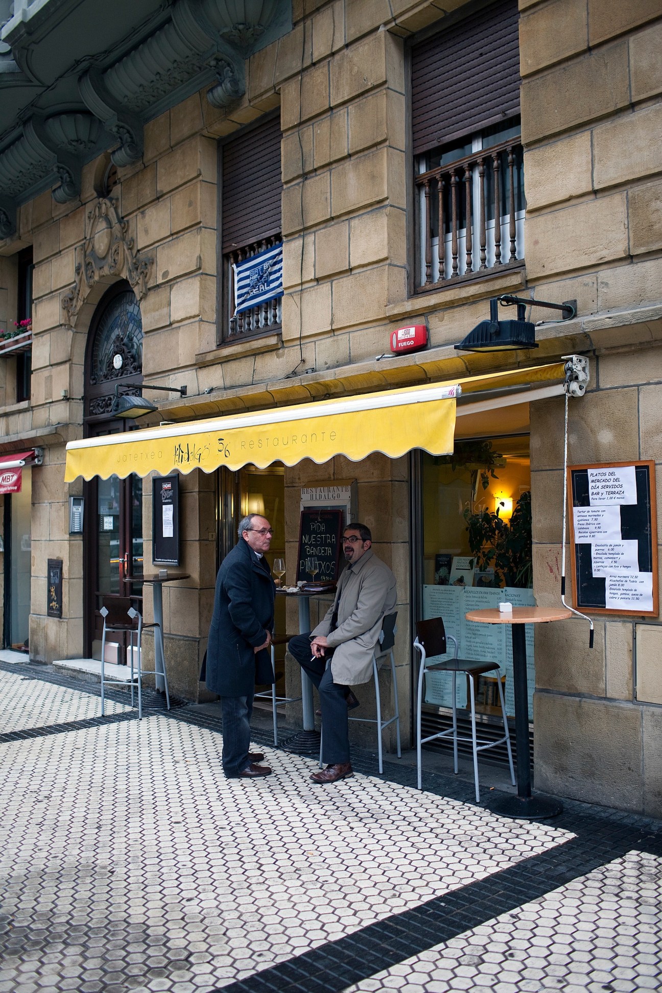 Exterior of Hidalgo 56 restaurant with yellow awning and two men standing outside.