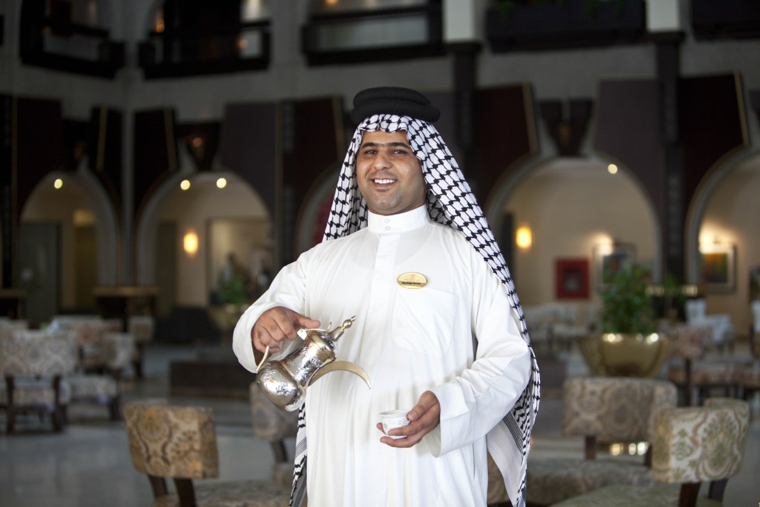 Man in traditional white dishdasha and black-and-white ghutra holding a decorative brass coffee pot in an arched courtyard.