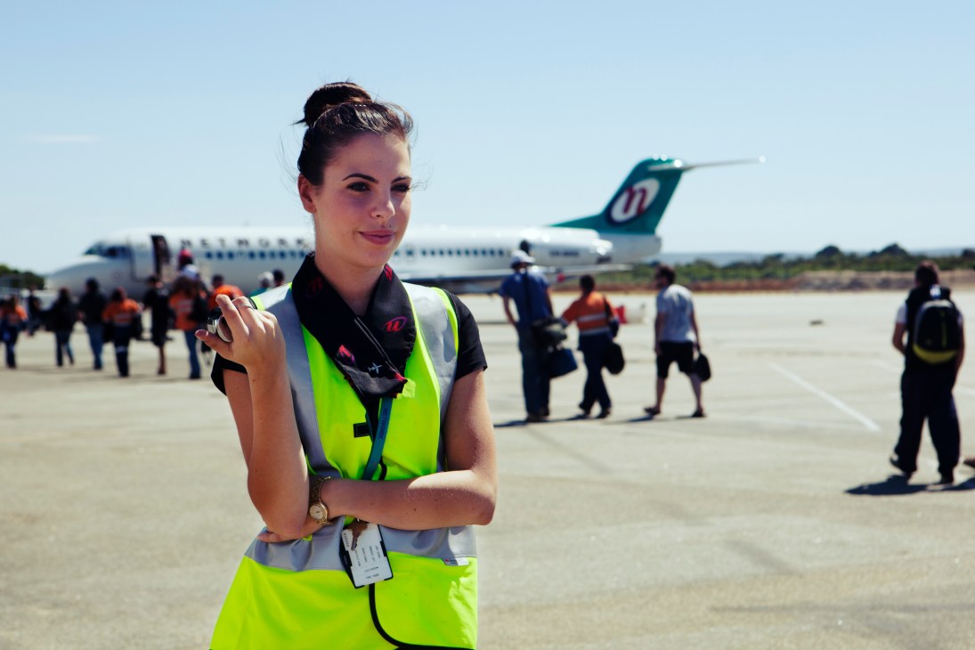Airport worker in high-visibility vest smiles at camera with aircraft and boarding passengers in background.