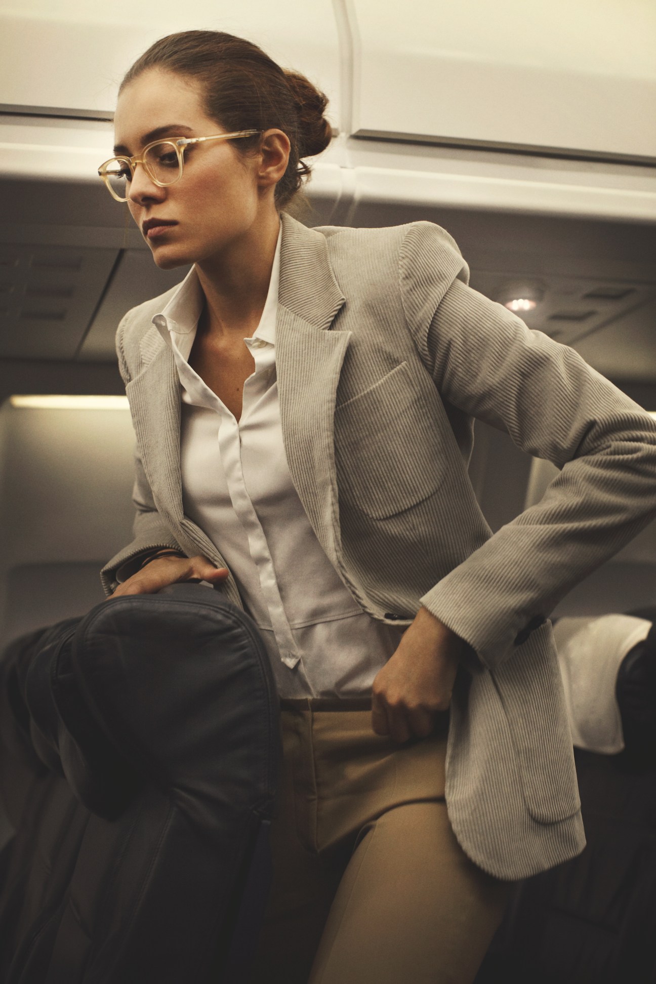 Woman wearing Oliver Peoples glasses, Trussardi jacket, Windsor shirt and Incotex trousers seated in aircraft cabin.