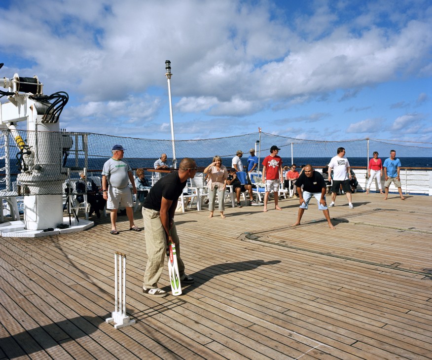 Cricket match on ship deck with officers and passengers playing while others watch near the railings.