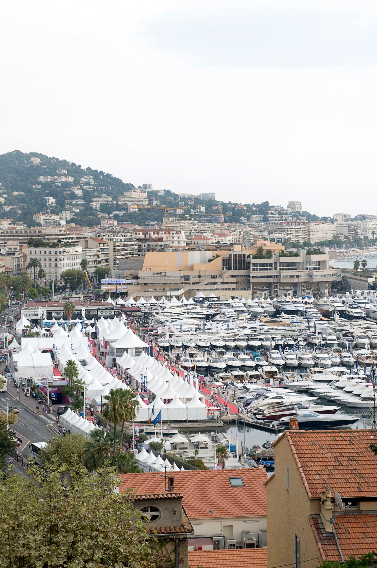 Aerial view of Cannes boat show with white tents, moored yachts, and hillside cityscape.