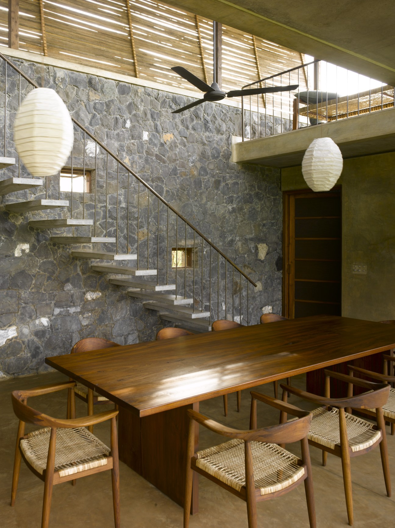Dining area with wooden table, mid-century chairs, black basalt stone wall, floating staircase and white paper lantern lights.
