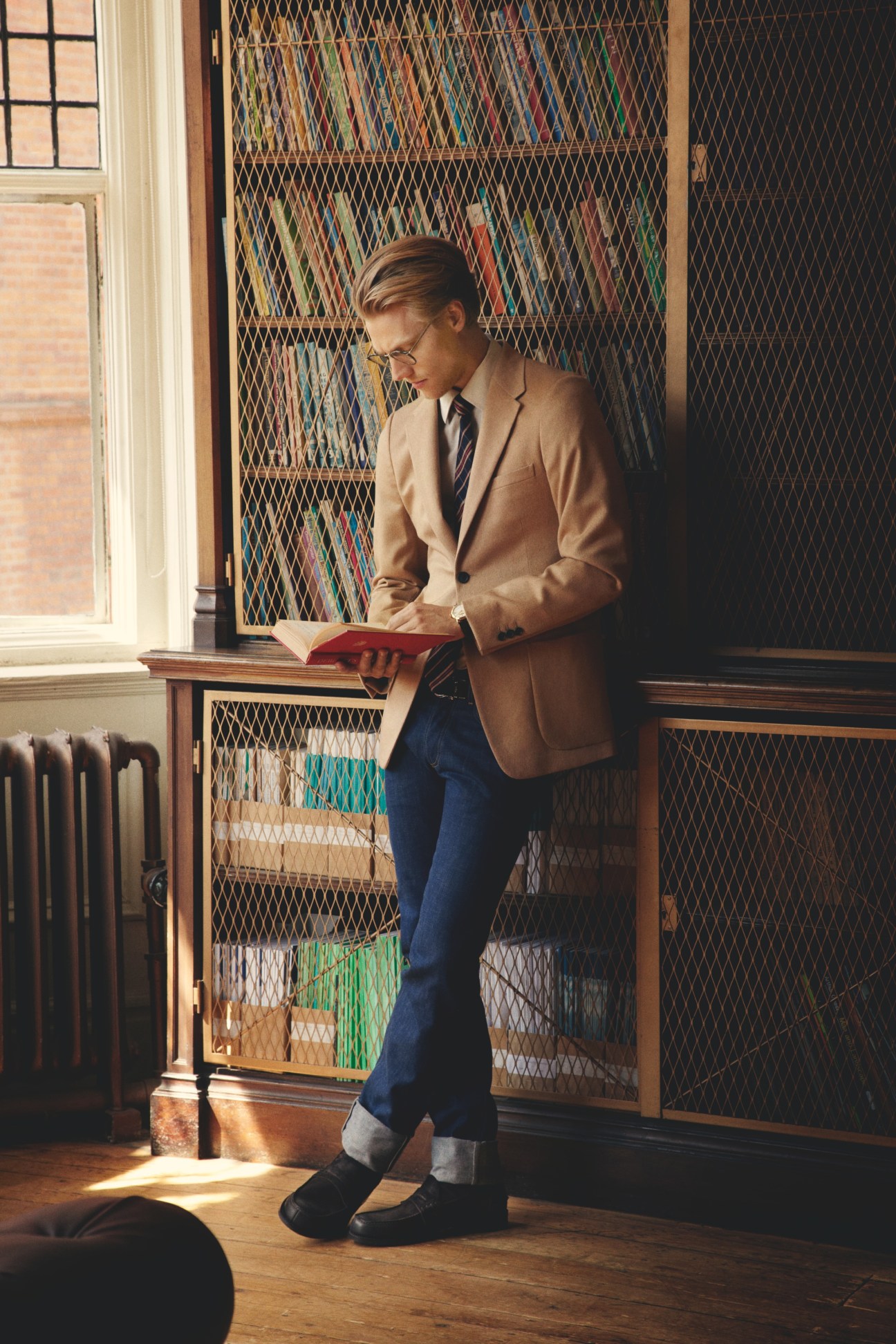 Man in Prada jacket, Bally shirt, and Gucci tie reading a red book by a library shelving unit.