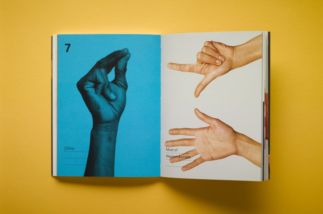 Open book showing hand gestures guide with blue page displaying Chinese hand gesture and white page showing Western European gestures against yellow background.