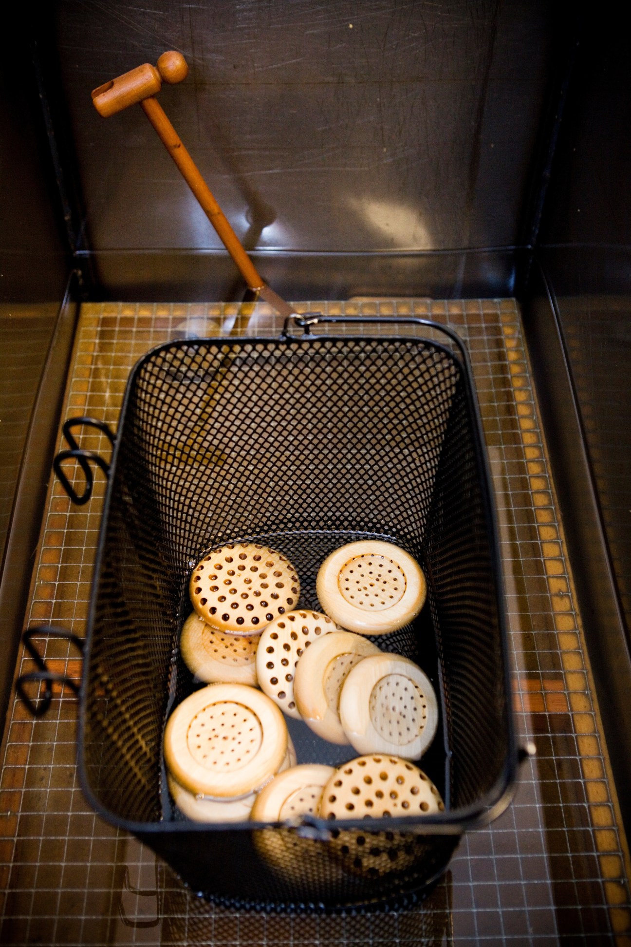 Wooden brush heads with perforated metal bases sit in a black mesh strainer next to a wooden-handled tool on a checkered surface.