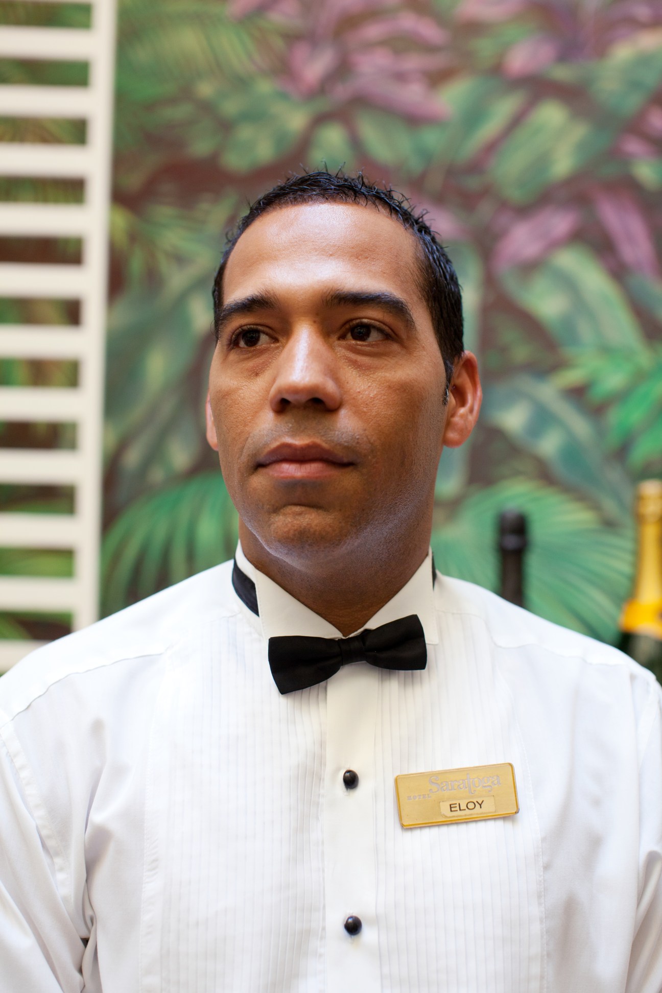 A waiter in formal white shirt and bow tie with name badge stands in front of tropical mural.
