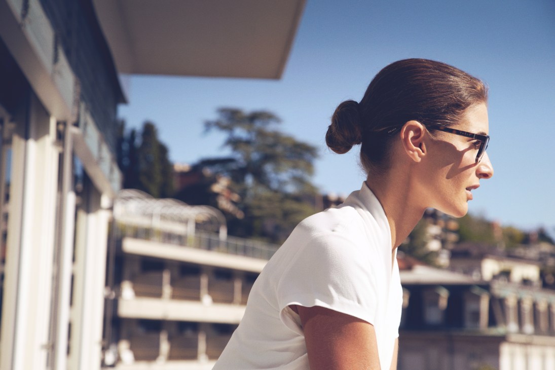 Woman wearing Prada glasses and Ralph Lauren dress overlooking Lugano lakeside buildings.