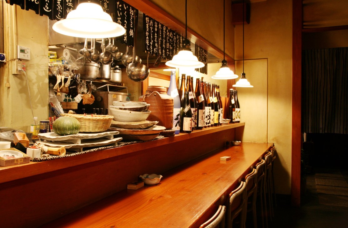 Counter seating at Ikedaya restaurant with kitchen visible behind, hanging cookware, pendant lights, and sake bottles on shelves.