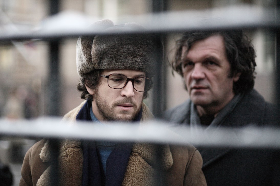 Two men in winter coats stand behind a metal railing in a cold urban setting.