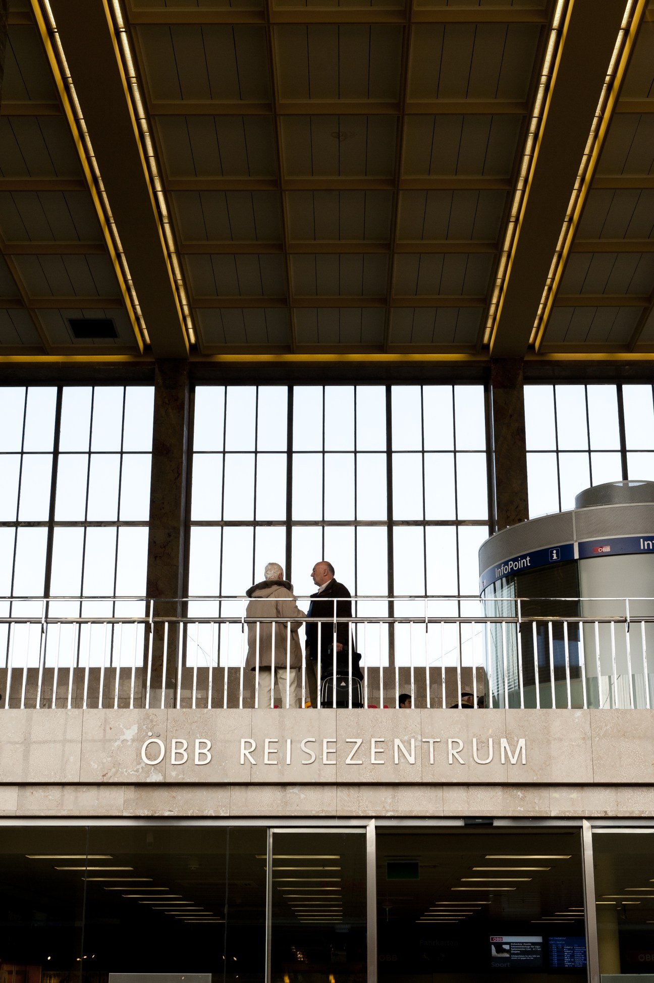 Two travelers converse on a modern railway station platform with gold-trimmed ceiling and large windows.