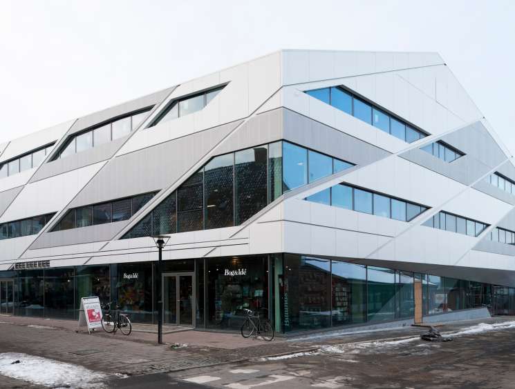 Modern white building with curved architectural lines and large windows, featuring ground-floor retail spaces and bicycles parked outside.
