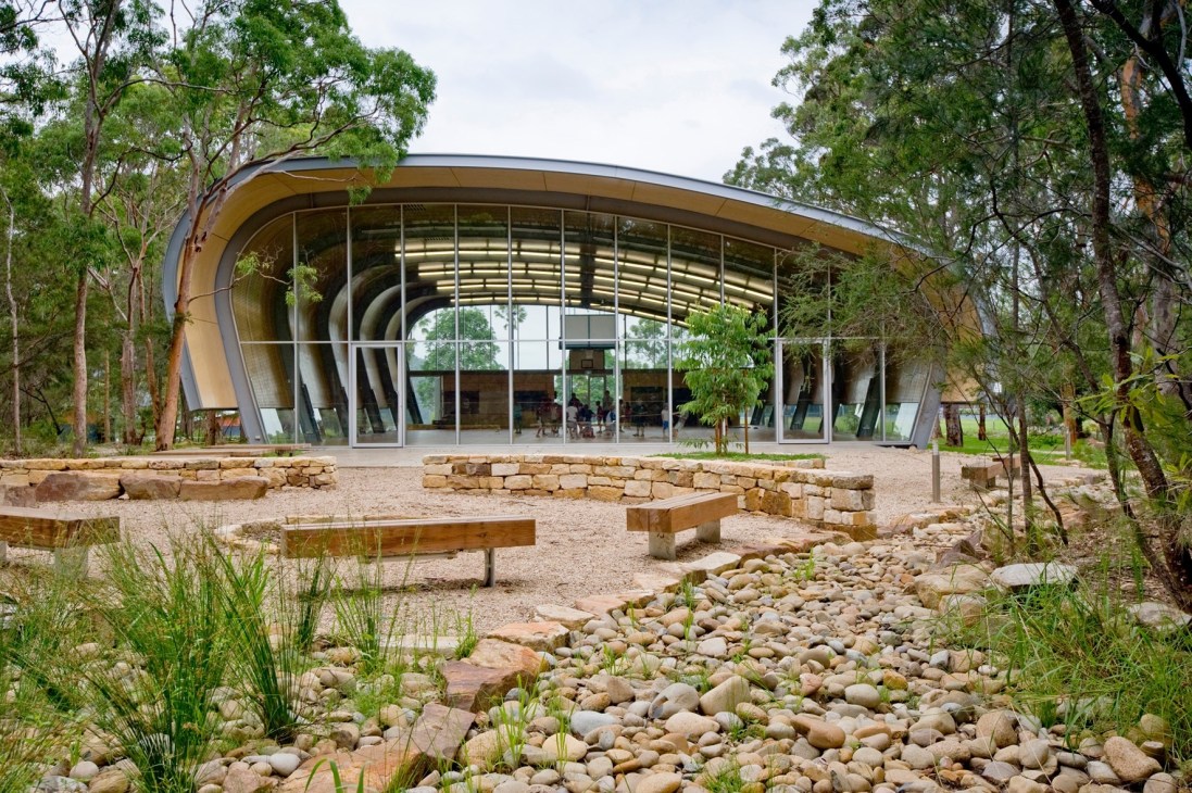 Curved modern sports hall with glass facade nestled among eucalyptus trees in Australian bushland.