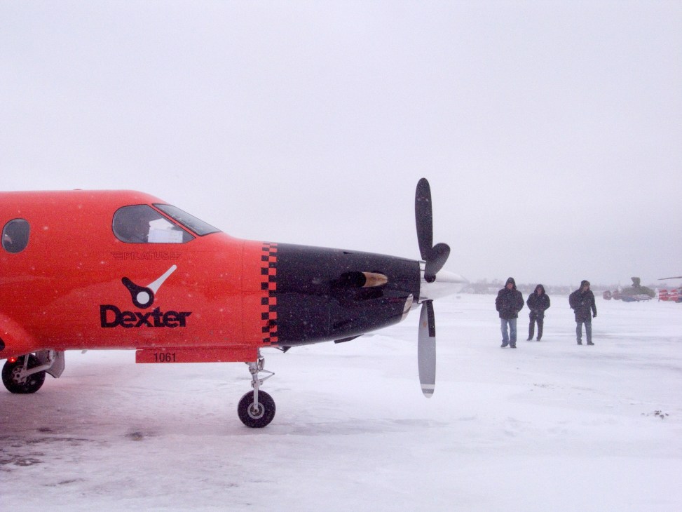 A bright orange and black Pilatus PC-12 turboprop aircraft branded with Dexter logo parked on snowy tarmac with people standing nearby.