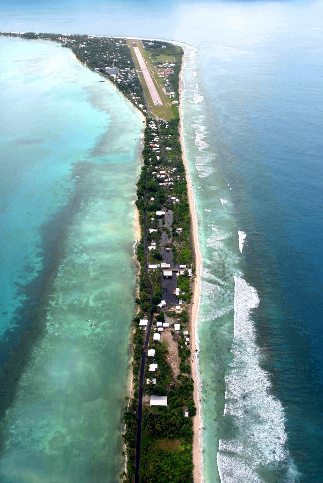 Aerial view of a narrow Pacific island with buildings, vegetation, and an airstrip surrounded by turquoise lagoon and ocean waves.