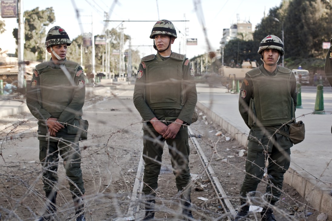 Three armed soldiers in military uniforms and helmets stand on a street behind barbed wire.