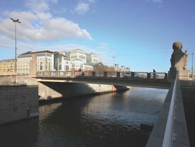 Brussels canal with modern waterfront development, bridge, and historic buildings under blue sky.