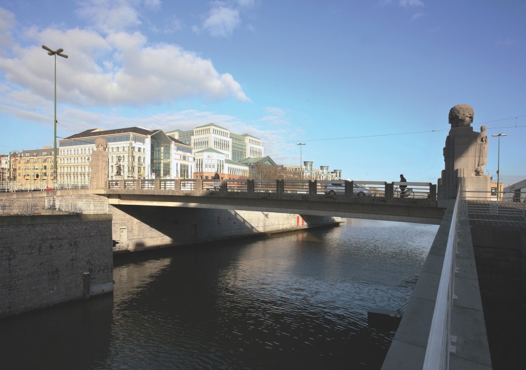 Brussels canal with modern waterfront development, bridge, and historic buildings under blue sky.